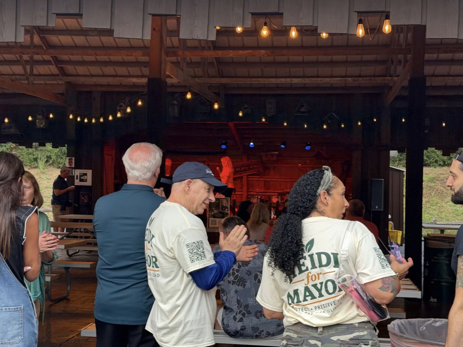 Volunteers in Heidi for Mayor shirts clap as Heidi speaks on the pavilion stage under blue spotlights.