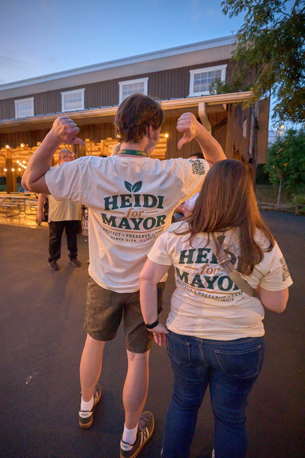 Two volunteers, seen from behind, point their thumbs at Heidi for Mayor printed on their shirt backs.